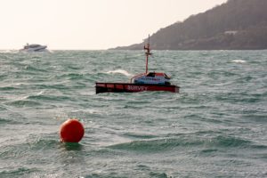 HydroSurv Uncrewed Surface Vessel (USV) on the sea with a bouy in the foreground and a boat and coastline in the background